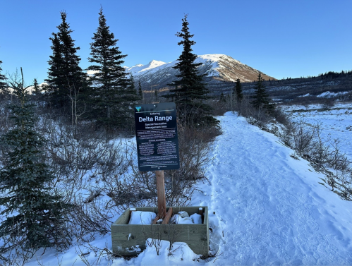 A snowy path leads to a sign about the Delta Range, surrounded by evergreen trees and mountains in the background.