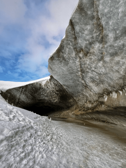 A close-up view of a glacier's icy surface, with a dramatic overhang and a blue sky in the background.