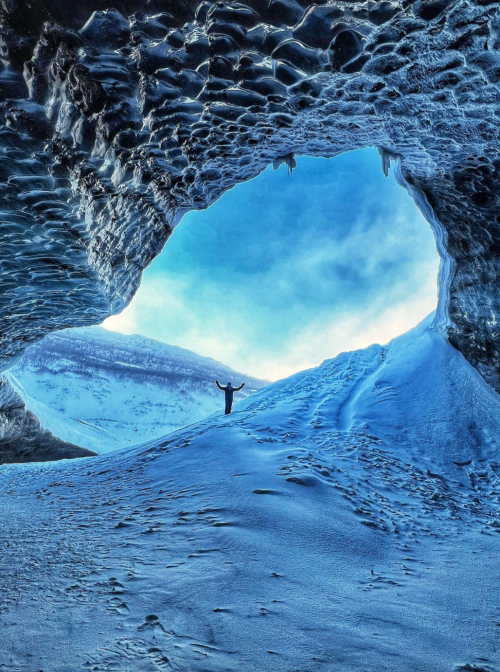 A person stands in an icy cave, surrounded by snow and ice formations, with a bright sky visible through the opening.