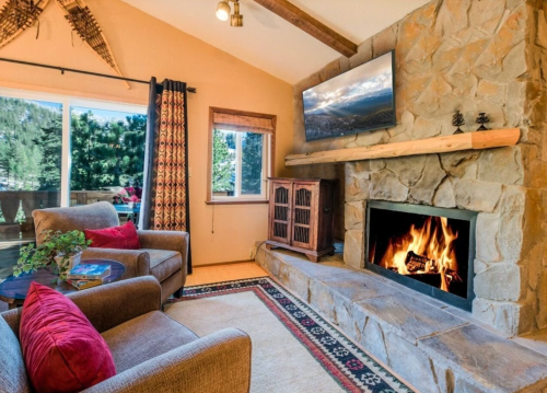 Cozy living room with a stone fireplace, comfortable chairs, and a large window showcasing a forest view.