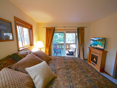 Cozy bedroom with a patterned bedspread, lamp, TV, and balcony view of trees. A fireplace adds warmth to the space.
