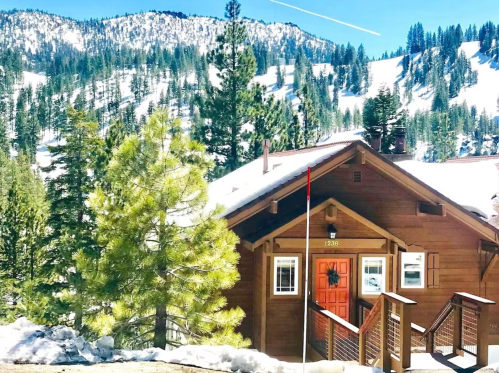 A cozy wooden cabin surrounded by snow-covered mountains and tall pine trees under a clear blue sky.