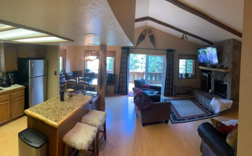Cozy living room and kitchen area with wooden beams, large windows, and a stone fireplace.