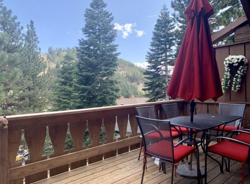A wooden balcony with a table and red umbrella, surrounded by tall pine trees and a scenic mountain view.