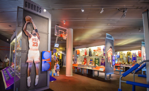 A sports museum display featuring basketball jerseys, memorabilia, and a hoop, with a player image on the wall.