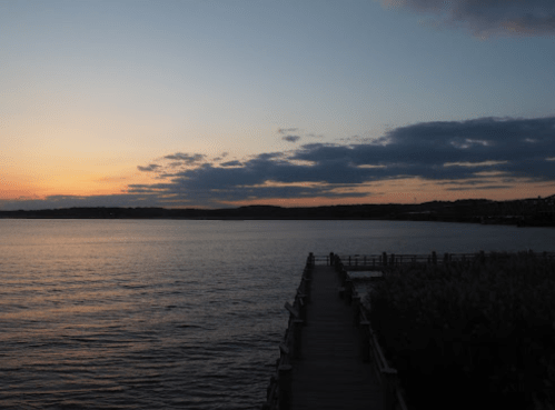 A serene sunset over a calm lake, with a wooden pier extending into the water and clouds in the sky.