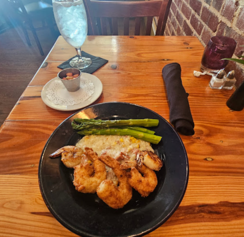 A plate of shrimp with asparagus and grits, accompanied by a glass of water and a small bowl of nuts on a wooden table.