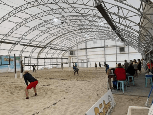 Indoor beach volleyball game in a large, covered sand court with spectators seated nearby.
