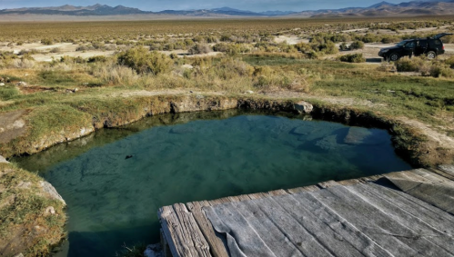 A natural hot spring surrounded by grasslands and mountains, with a wooden platform and a parked car nearby.
