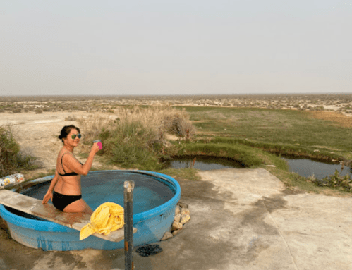 A woman in a bikini relaxes in a blue hot tub, surrounded by a scenic landscape and hazy sky.