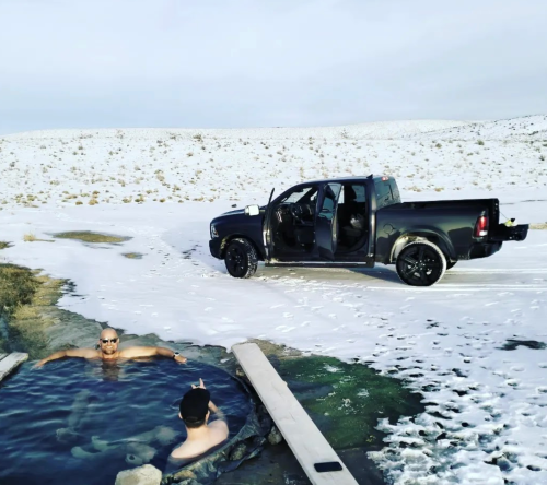 Two people relax in a hot tub surrounded by snow, with a black pickup truck parked nearby.