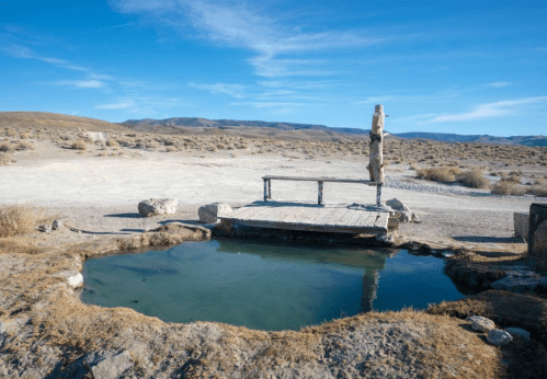 A natural hot spring surrounded by dry landscape and a wooden bench under a clear blue sky.