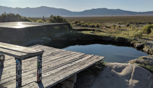 A wooden platform beside a natural hot spring, with mountains and a vast landscape in the background under clear skies.