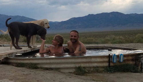 A man and a woman relax in a metal tub outdoors, with two dogs nearby and mountains in the background.