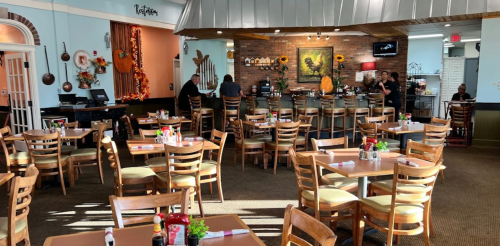 A cozy restaurant interior with wooden tables, chairs, and a bar area, decorated for fall. Staff are serving customers.