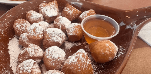 A plate of powdered sugar-dusted beignets with a small cup of syrup on the side.