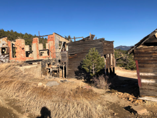 Abandoned buildings in a rural area, surrounded by dry grass and trees, under a clear blue sky.