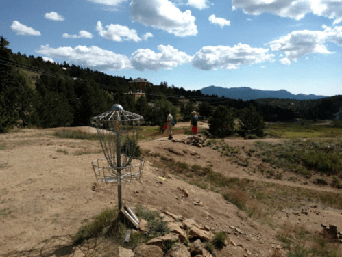 A disc golf basket in the foreground with two players walking on a dirt path in a mountainous landscape under a blue sky.