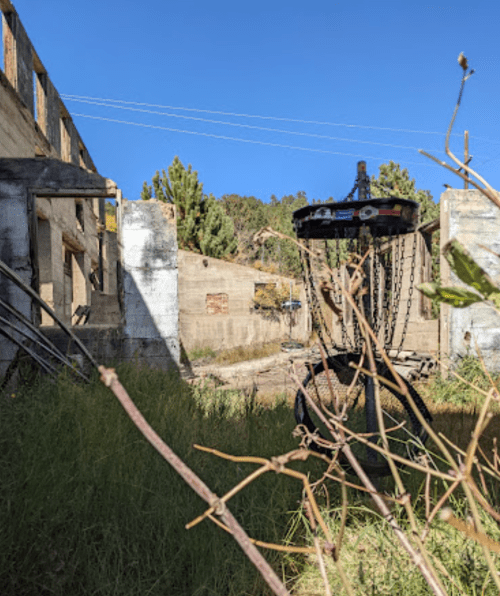 An abandoned building with a disc golf basket in a grassy area, surrounded by trees and blue sky.