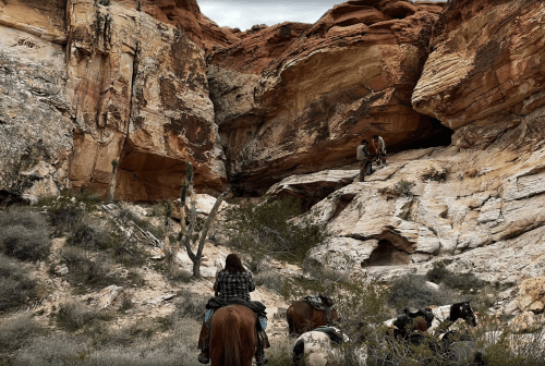 A person on horseback watches two people exploring a rocky canyon landscape with red rock formations.