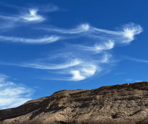 Wispy clouds swirl above a rugged mountain landscape under a clear blue sky.