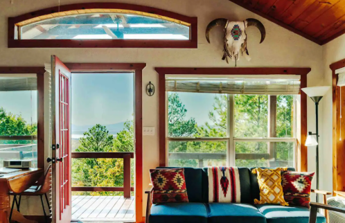 Cozy living room with a blue couch, colorful pillows, large windows, and a view of trees and mountains outside.