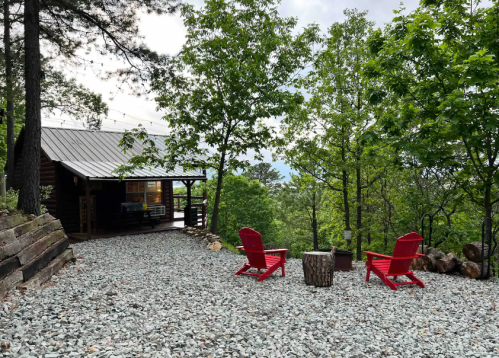 A cozy cabin surrounded by trees, featuring two red chairs and a small table on a gravel path.