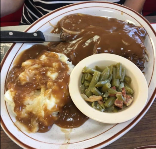 A plate of meat with brown gravy, mashed potatoes, and a side of green beans in a bowl.