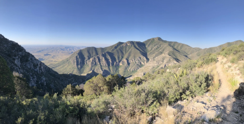 Panoramic view of rugged mountains and valleys under a clear blue sky, with lush greenery in the foreground.