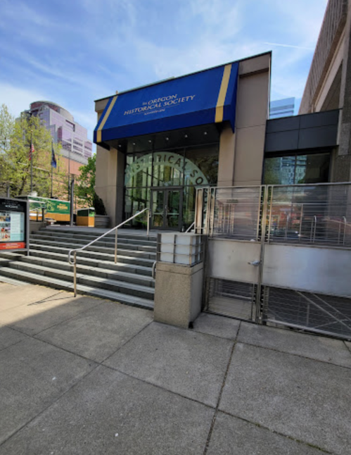 Entrance of the Oregon Historical Society, featuring steps and a blue awning, with city buildings in the background.