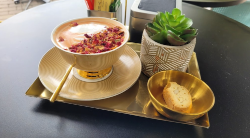 A decorative cup of rose-infused tea with petals, a small cookie, and a succulent plant on a gold tray.