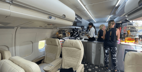 Interior of a private jet with a galley area where three women are preparing food and drinks. Comfortable seating visible.