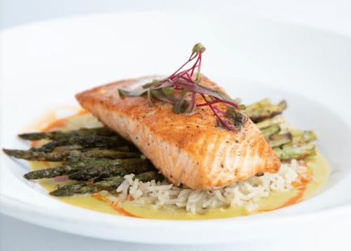 Grilled salmon fillet on a bed of rice and asparagus, garnished with microgreens and a drizzle of sauce.