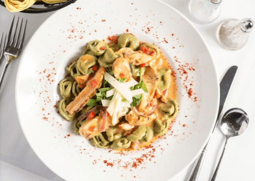 A plate of creamy tortellini with chicken, red peppers, and garnished with cheese and herbs on a white tablecloth.