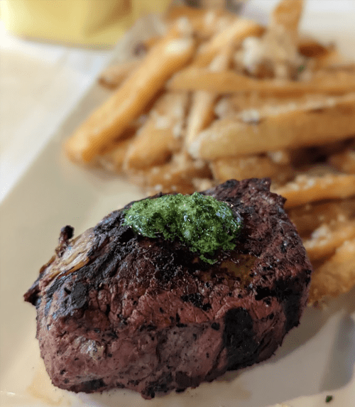 A juicy steak topped with green sauce, served alongside crispy French fries on a white plate.