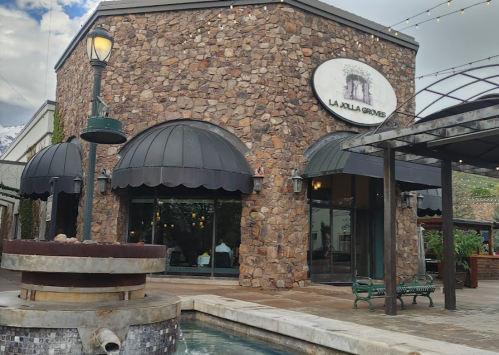 Exterior of La Jolla Groves restaurant featuring stone walls, black awnings, and a fountain in front.