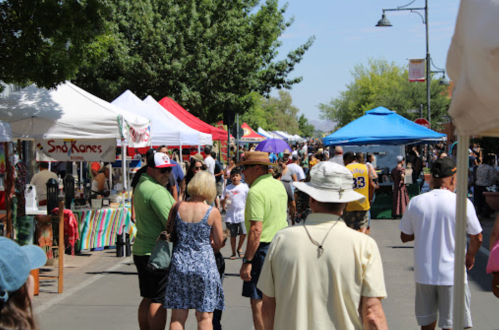 A bustling outdoor market with colorful tents, people walking, and trees lining the street on a sunny day.