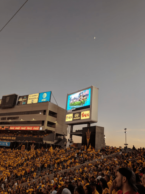 A crowded stadium at sunset, with a large screen displaying a sports event and a crescent moon in the sky.