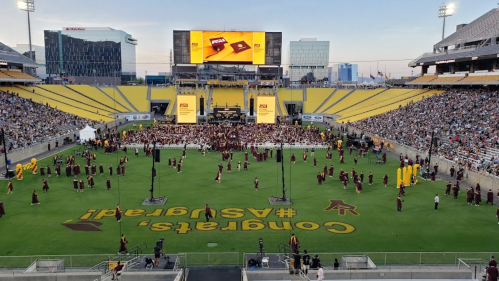 A large graduation ceremony at a stadium, with graduates in maroon gowns and a crowd watching from the stands.