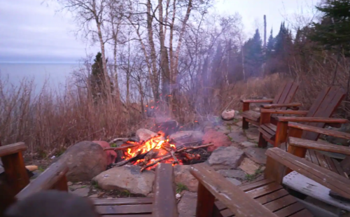 A cozy campfire surrounded by wooden chairs, with trees and a lake in the background at dusk.