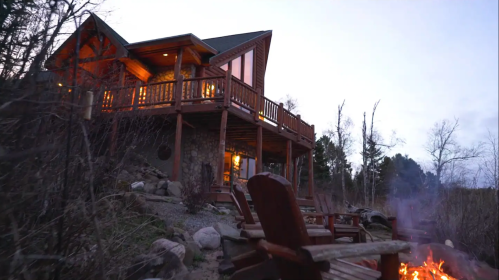 A cozy cabin with a wooden deck, surrounded by trees and rocks, with a fire pit in the foreground at dusk.