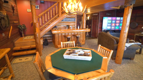 Cozy cabin interior featuring a wooden bar, dining table, and rustic decor with a staircase and large TV.