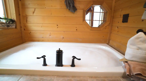 A cozy bathroom featuring a large, white soaking tub with black faucets, surrounded by wooden walls and a window.
