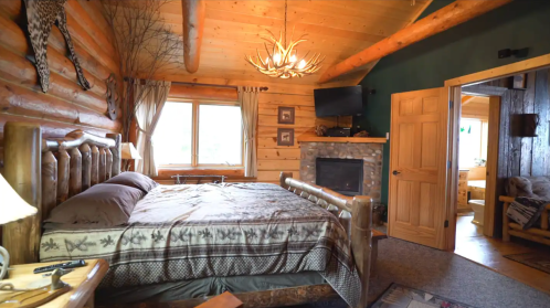 Cozy log cabin bedroom with a wooden bed, stone fireplace, and large window, featuring rustic decor and warm lighting.