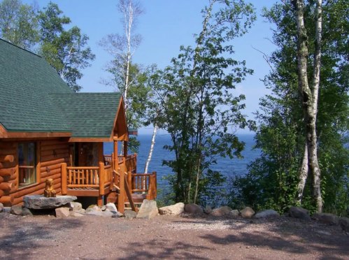 A cozy log cabin surrounded by trees, overlooking a serene lake under a clear blue sky.