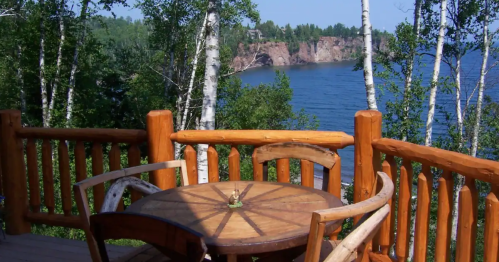 A wooden deck with a round table and chairs overlooks a serene lake surrounded by trees and cliffs.
