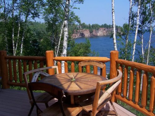 A wooden table and chairs on a deck overlooking a serene lake and rocky cliffs, surrounded by lush trees.