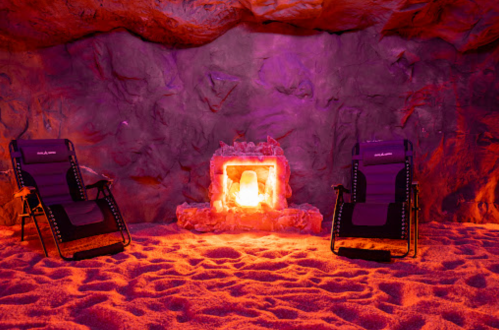 Two lounge chairs in a cozy cave setting with a glowing fireplace and purple lighting, surrounded by sandy ground.