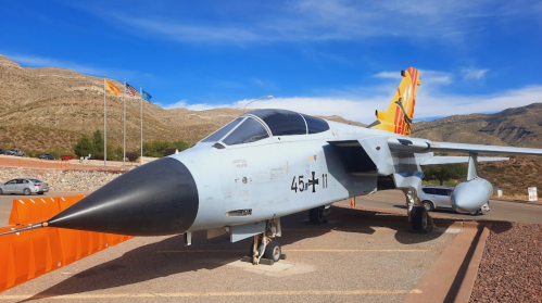 A military jet displayed outdoors, with a mountainous backdrop and clear blue sky.