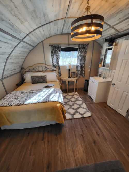 Cozy bedroom with a curved ceiling, a bed, nightstand, and decorative window curtains. Warm lighting and wooden floors.